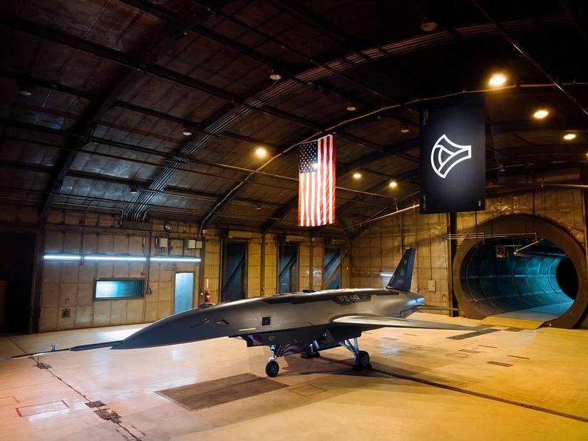 A photo of the Anduril Fury drone in a dimly lit hangar with both the American and Aduril flags hanging on the ceiling.
