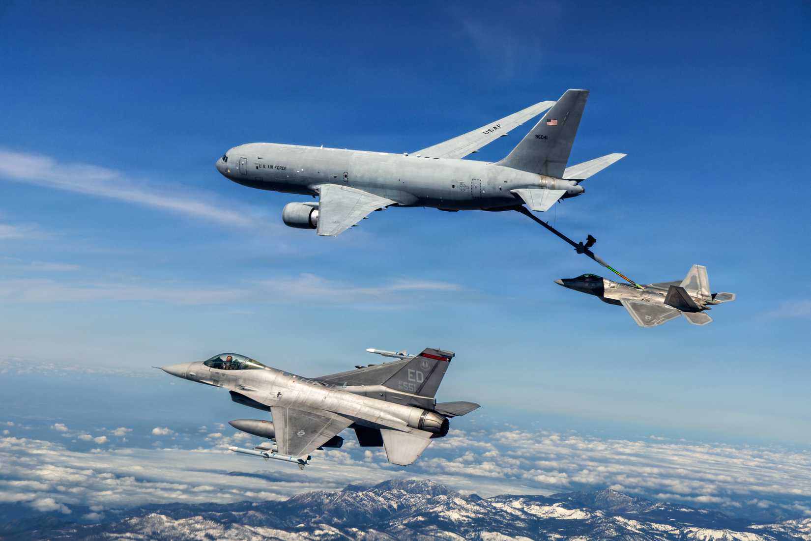 A photo of a KC-46 Pegasus refueling a F-22 Raptor while an F-16 is in the foreground, above a mountain range.