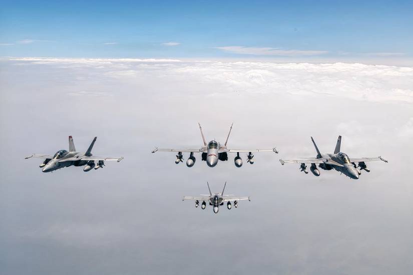 A head-on photo showing four F/A-18s flying above a sheet of grey clouds. 