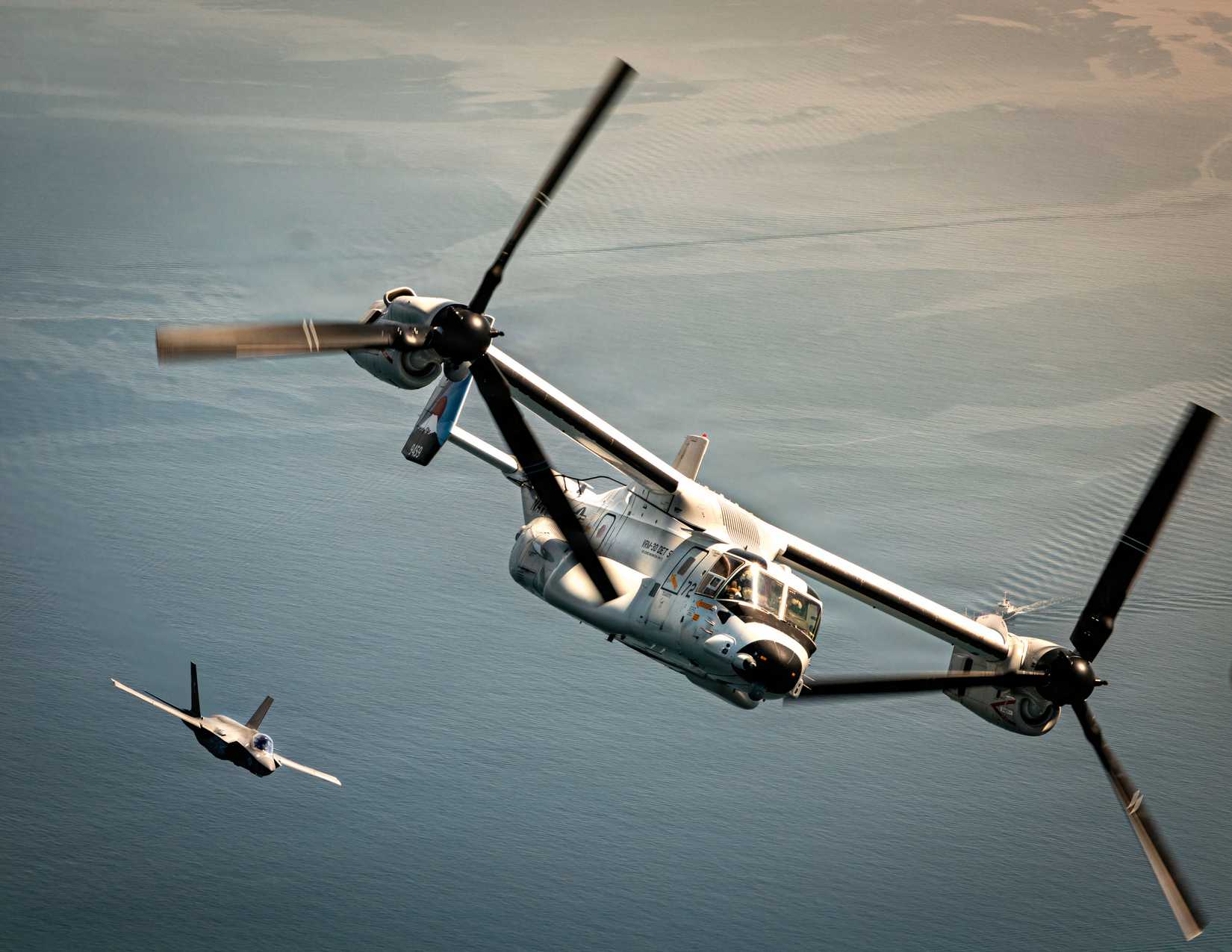 A photo showing a V-22 Osprey in the foreground with an F-35 close behind, they are flying over water. 