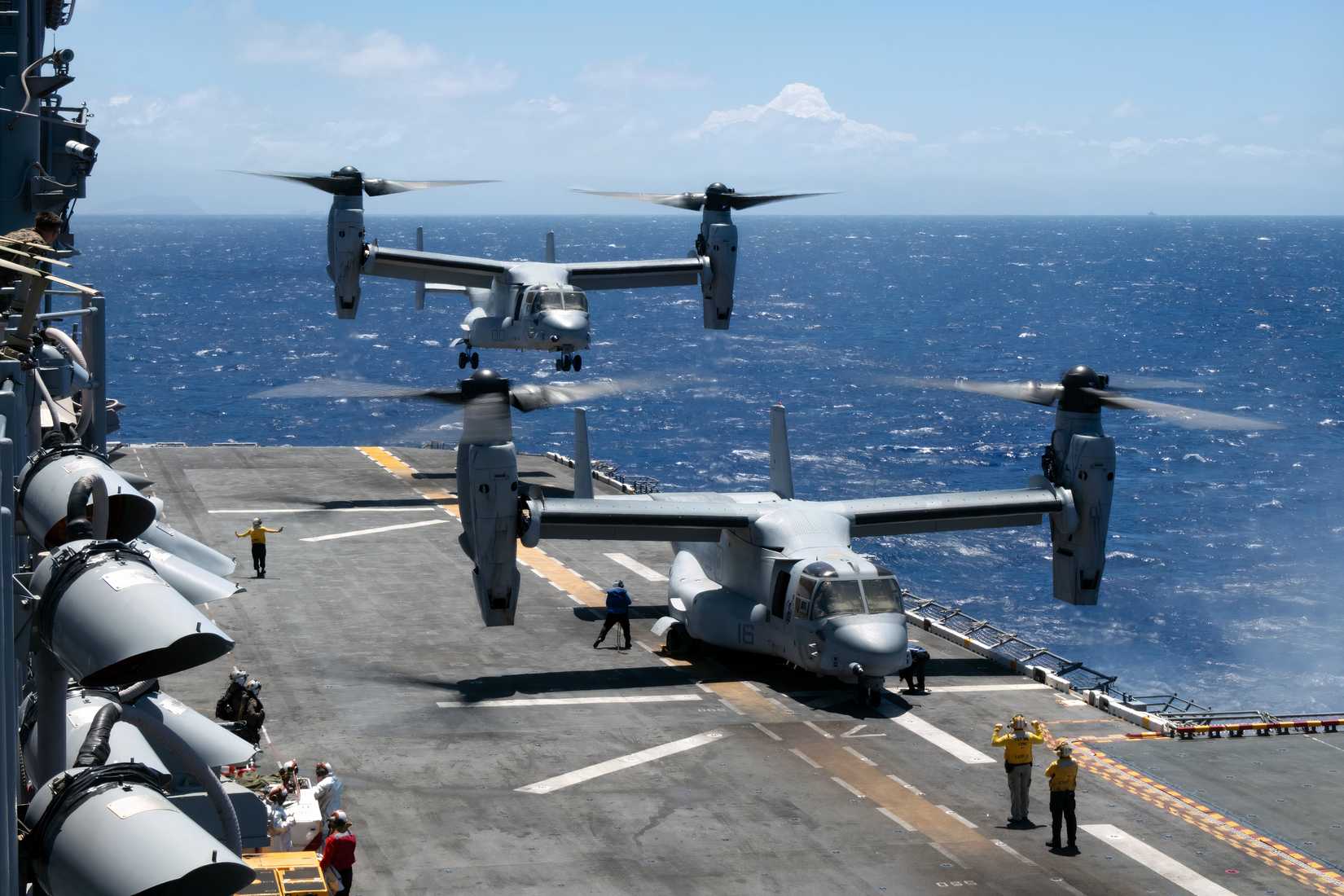 A photo showing two V-22 Osprey's on the flight deck of an amphibious assault ship. One has just lifted off, the other is about to do the same. 