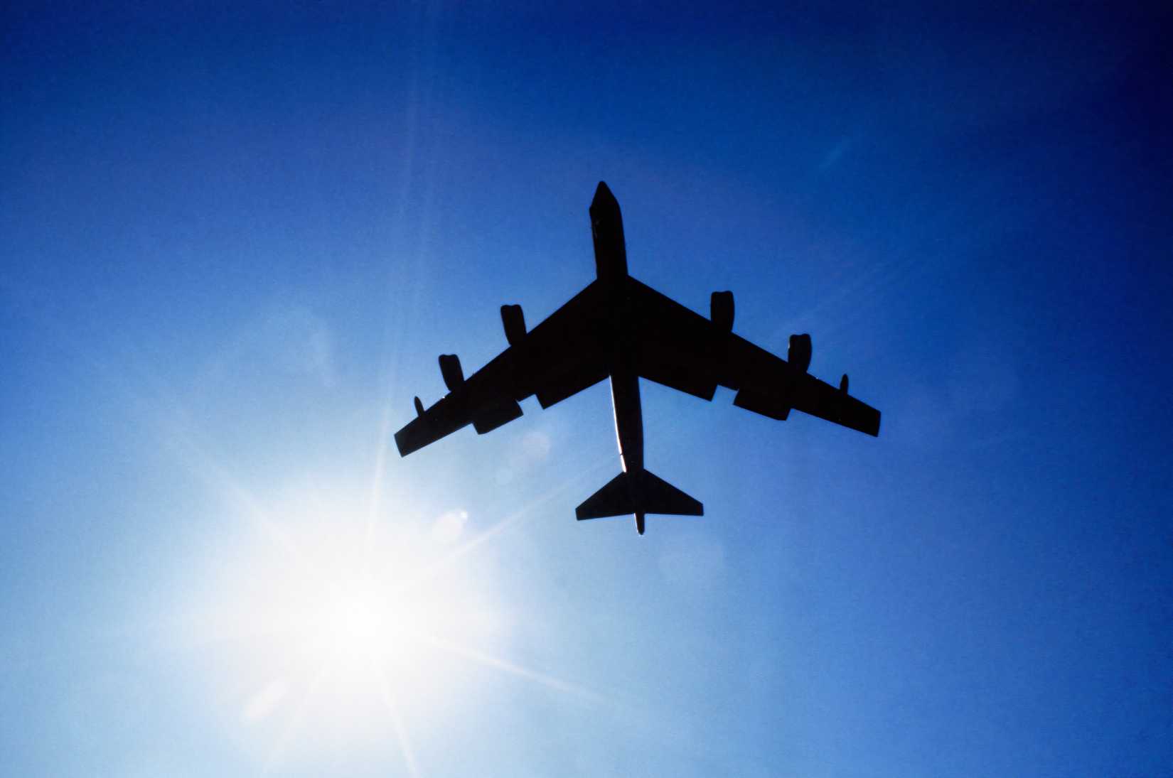 A photo of a B-52's silhouette against a blue sky with the sun shinning near the left wing tip.