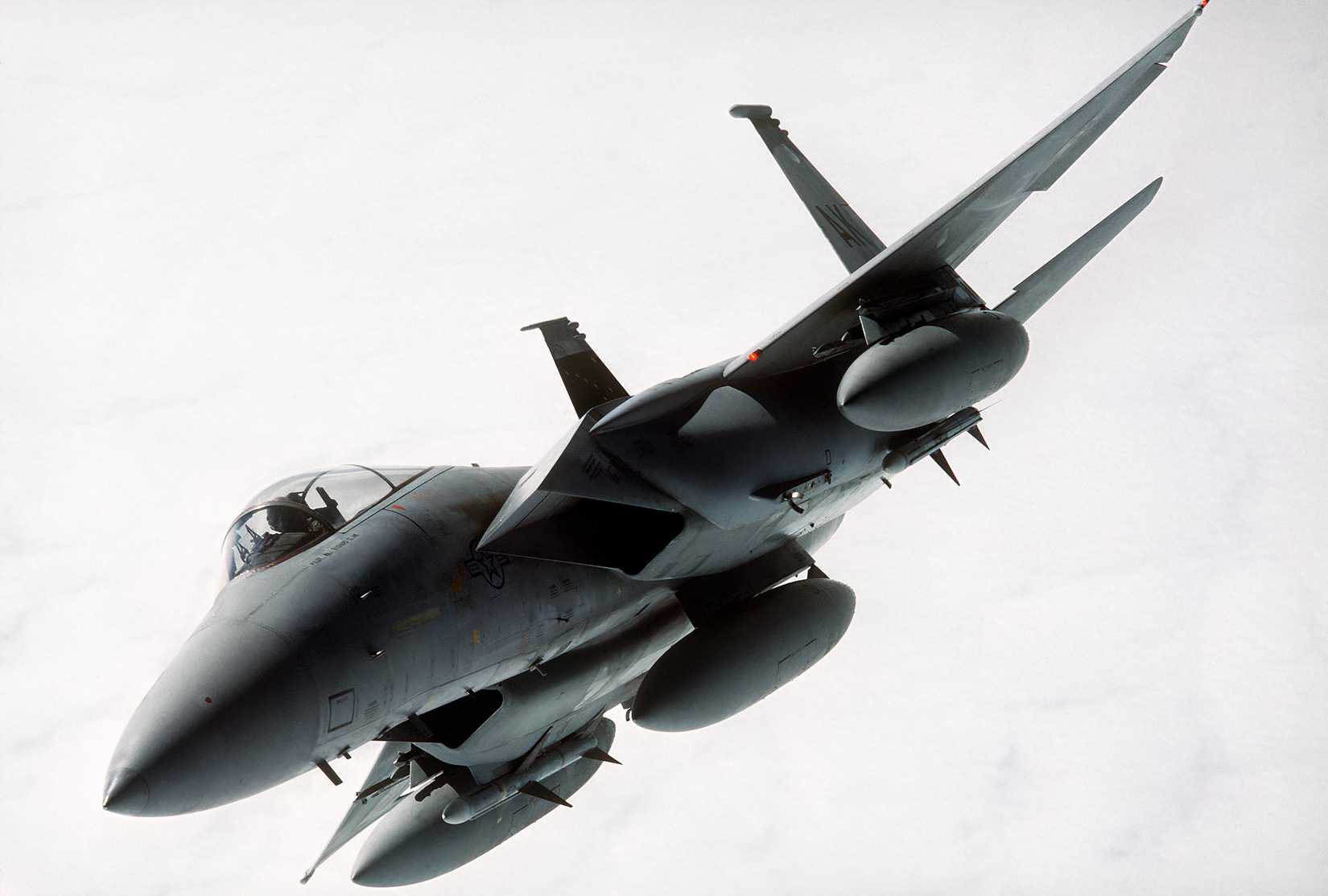 A photo of an F-15 Eagle against a wall of grey clouds, making a turn showing its undercarriage, and two missiles.