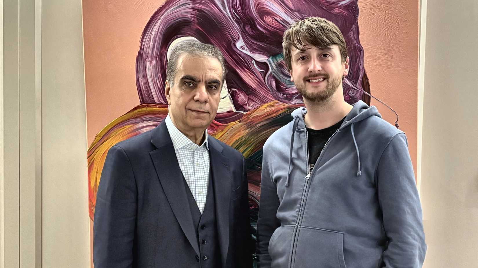 Air Arabia Group CEO Adel Al-Ali standing alongside Simple Flying Content Director Tom Boon. Adel Al-Ali is dressed in a dark three-piece suit with a patterned dress shirt, while Tom Boon wears a casual blue zip-up hoodie, both posing together in front of a colorful, abstract painted artwork featuring swirls of purple, yellow, and orange tones. 