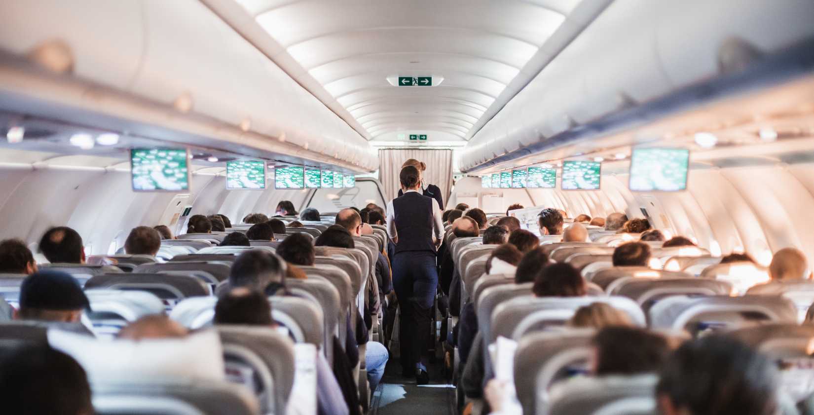 Interior of airplane with passengers on seats and stewardess in uniform walking the aisle, serving people. Commercial economy flight service concept