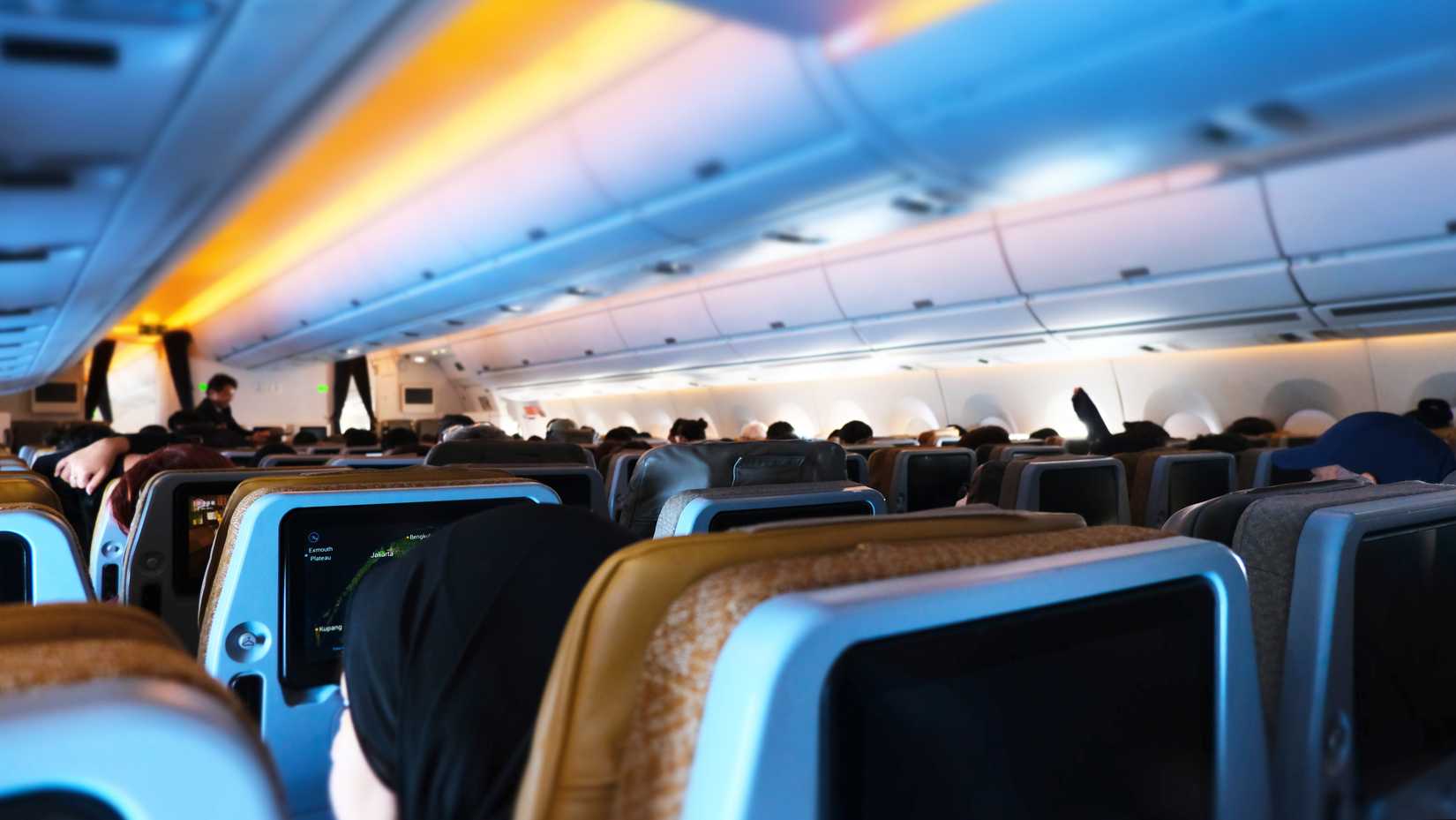 view of economy class cabin interior with passengers seated inside of Singapore Airlines Airbus type airplane with blue and yellow ambient lightings. airplane interior