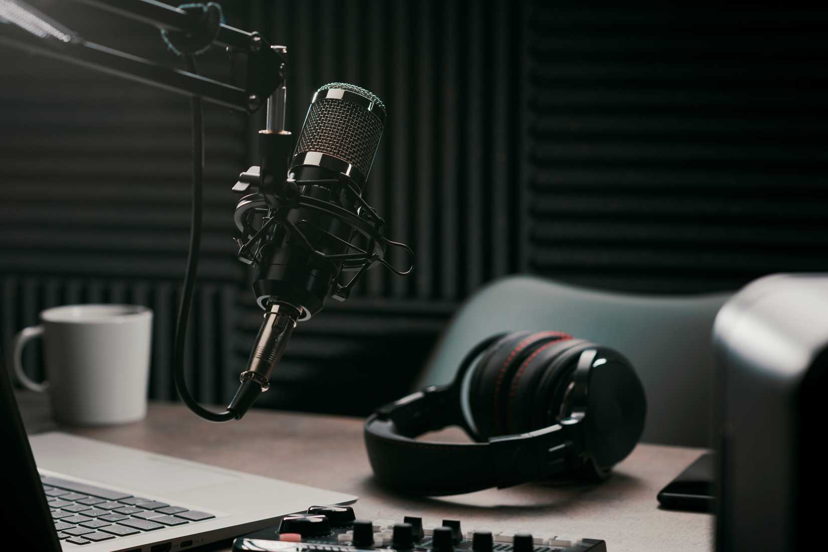 A suspended microphone, headphones, laptop and audio controls in a dark recording room, representing podcasting, radio, voiceover, narration and training for spoken communication.