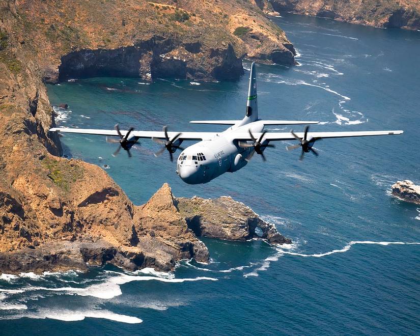 A C-130 flying over water and rocky outcroppings. 
