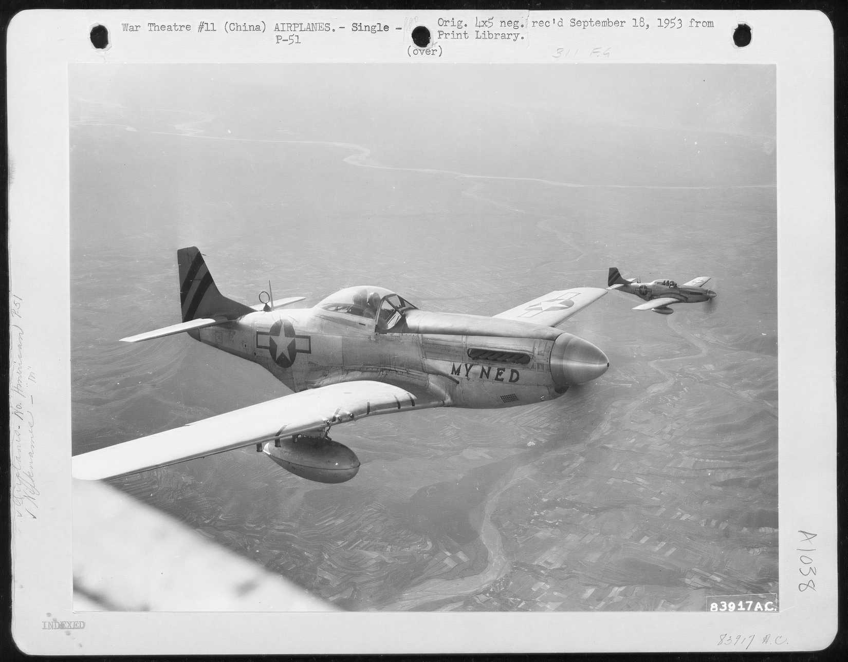 A black and white photo of two P-51 Mustangs flying over a rural area below